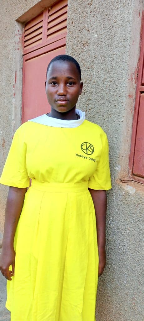 Nayiga Daisy Babirye, a young girl in a bright yellow dress, standing outdoors in front of a textured wall, representing hope and education support through Pearl Global Ministries.