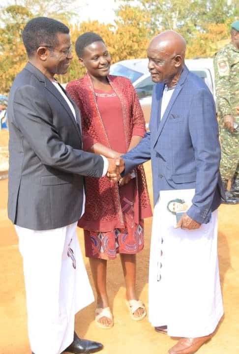 Three individuals shaking hands outdoors during a community event, with two men in traditional attire and a woman in a red dress, in front of a vehicle and trees.