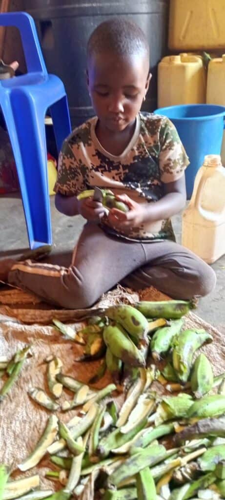 Young boy sitting on the floor, sorting fresh green beans with a focused expression, surrounded by cleaning tools and containers in a community setting.